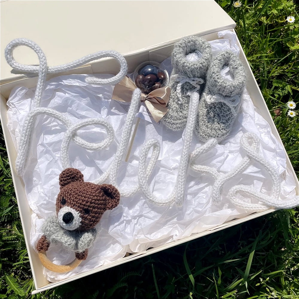 Zestaw prezentowy dla noworodka, z ręcznie wykonywanymi elementami. W zestawie znajduje się: imię z włóczki, skarpetki/buciki, grzechotka w kształcę misia i liofilizowane maliny w czekoladzie w formie lizaka.